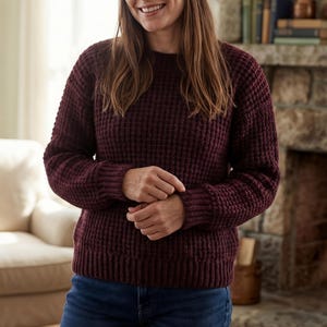 May include: A burgundy-colored, waffle-knit sweater with a crew neck and long sleeves. The sweater has a textured appearance and a ribbed hem and cuffs. The person is wearing blue jeans. The sweater is a cozy, casual garment.