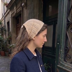 Peut inclure: Un foulard en tricot beige présentant un motif ajouré délicat avec un bord festonné. Le foulard est noué à l'arrière de la tête, mettant en valeur une construction en fil léger et texturé qui couvre le sommet du crâne tout en laissant les cheveux libres.