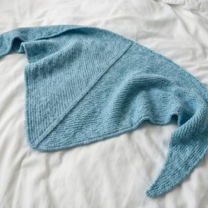 Puede incluir: Un chal tejido de color azul claro con forma triangular. El chal está hecho de hilo suave y tiene un aspecto texturizado. El chal está sobre una sábana blanca.