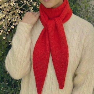 Puede incluir: Una bufanda roja brillante de punto se lleva sobre un suéter color crema. La bufanda está anudada en el cuello y cuelga en dos extremos largos y puntiagudos. El suéter tiene un diseño de punto trenzado.