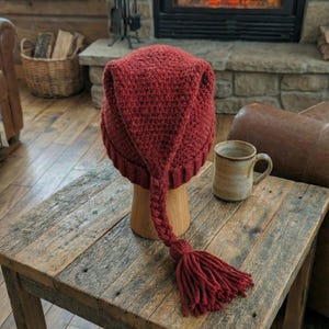 May include: A burgundy-colored, knitted hat with a long, braided tassel. The hat is displayed on a wooden stand atop a rustic wooden table. A ceramic mug sits nearby. The background features a fireplace with a burning fire.