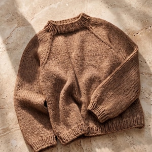 Könnte beinhalten: Ein langärmeliger, hellbrauner Strickpullover mit geripptem Rundhalsausschnitt, gerippten Bündchen und einem gerippten Saum. Das Kleidungsstück ist aus grobem Garn gefertigt und verfügt über Raglanärmel in einer durchgehend einheitlichen Farbe.