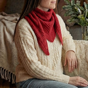 May include: A red knitted triangle scarf is draped around the neck of a person wearing a cream-colored cable knit sweater. The scarf has a textured pattern and is made of a soft, warm material. The sweater has a classic cable knit design.
