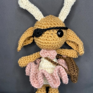 May include: A crocheted toy with brown fur, white horns, and a pink dress. The toy has a black eye patch and a brown bag.