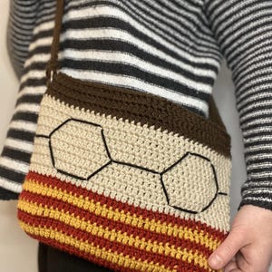 May include: A crocheted crossbody bag with a brown, beige, and red striped pattern. The bag features a honeycomb design in the center.