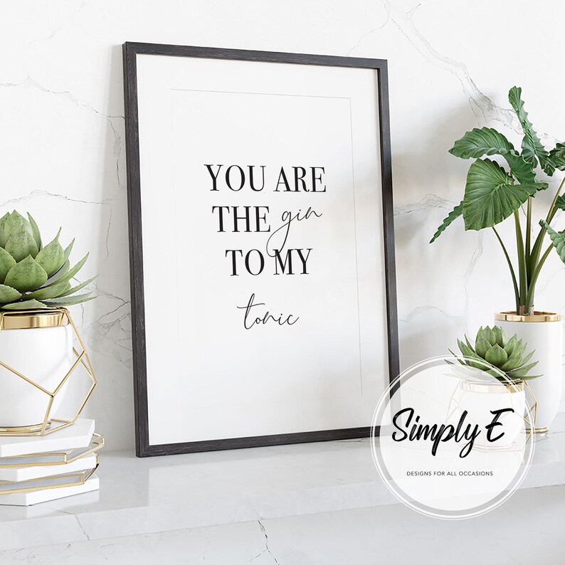 Puede incluir: Impresi&oacute;n enmarcada con el texto "YOU ARE THE gin TO MY tonic" en escritura negra. La impresi&oacute;n est&aacute; en un marco negro. Plantas decorativas en macetas blancas y doradas a ambos lados de la impresi&oacute;n.