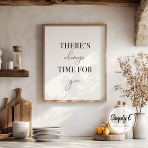 Puede incluir: Una impresi&oacute;n enmarcada con el texto "THERE'S always TIME FOR gin" en escritura negra. La impresi&oacute;n est&aacute; en un marco de madera clara, exhibida en un entorno de cocina con una paleta de colores neutros. Otros art&iacute;culos de cocina incluyen tablas de cortar, platos y un jarr&oacute;n con flores secas.