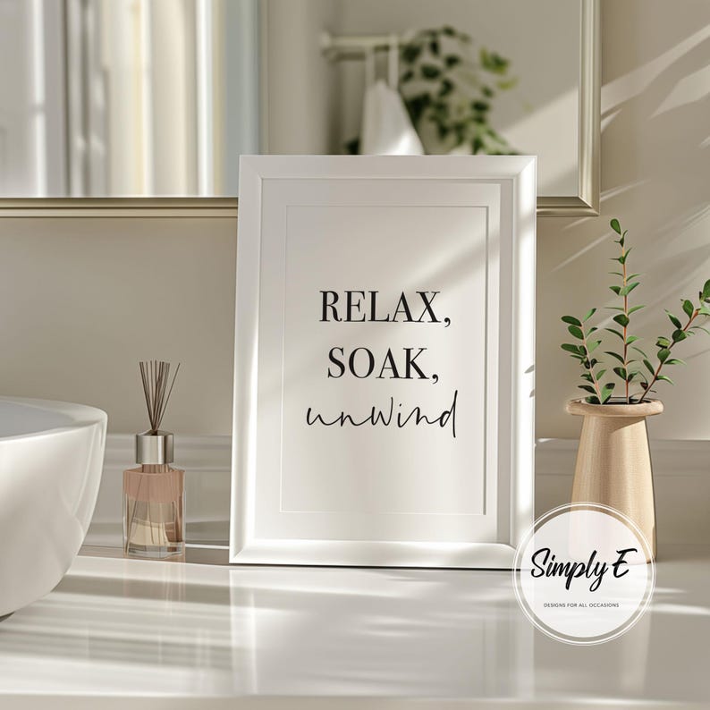 May include: A white framed print with the words "RELAX, SOAK, unwind" in black script. The print is displayed on a white surface, next to a small vase with greenery and a diffuser bottle. The image has a spa or bathroom theme.