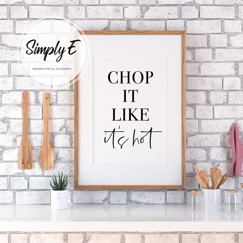 May include: A framed kitchen print with the text "Chop it like it's hot" in black lettering on a white background. The print is hanging on a white brick wall.