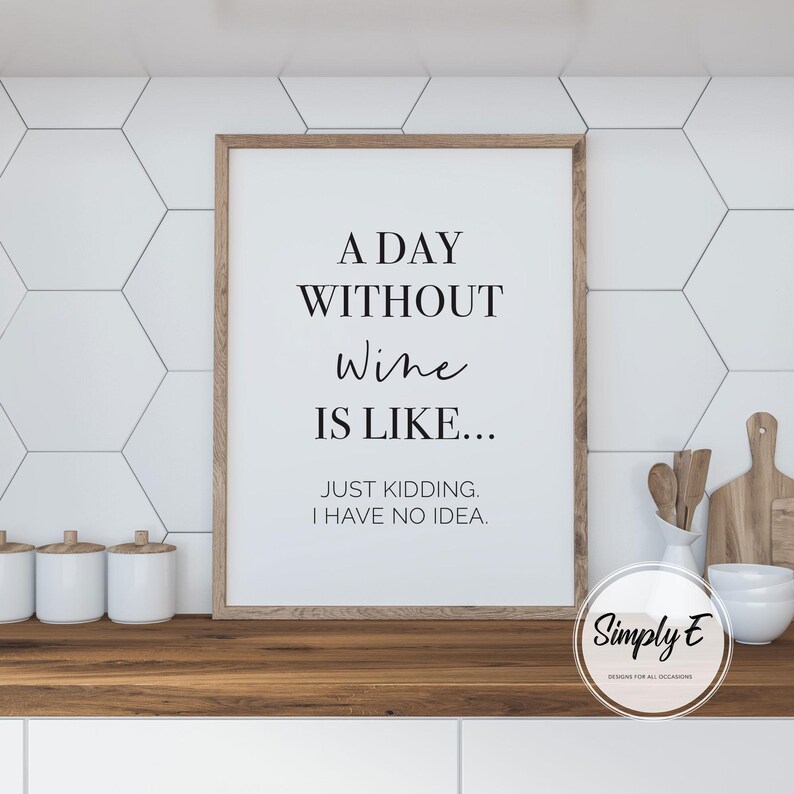 May include: A framed print with the text "A DAY WITHOUT Wine IS LIKE... JUST KIDDING. I HAVE NO IDEA." The print has a wooden frame and is displayed on a wooden countertop. The background features white hexagonal tiles.