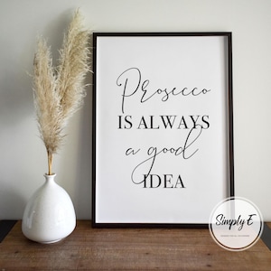 Op de afbeelding: Zwart-witte printbare wandkunst met de tekst "Prosecco is always a good idea". De print is ingelijst in een zwart frame en ligt op een houten oppervlak.