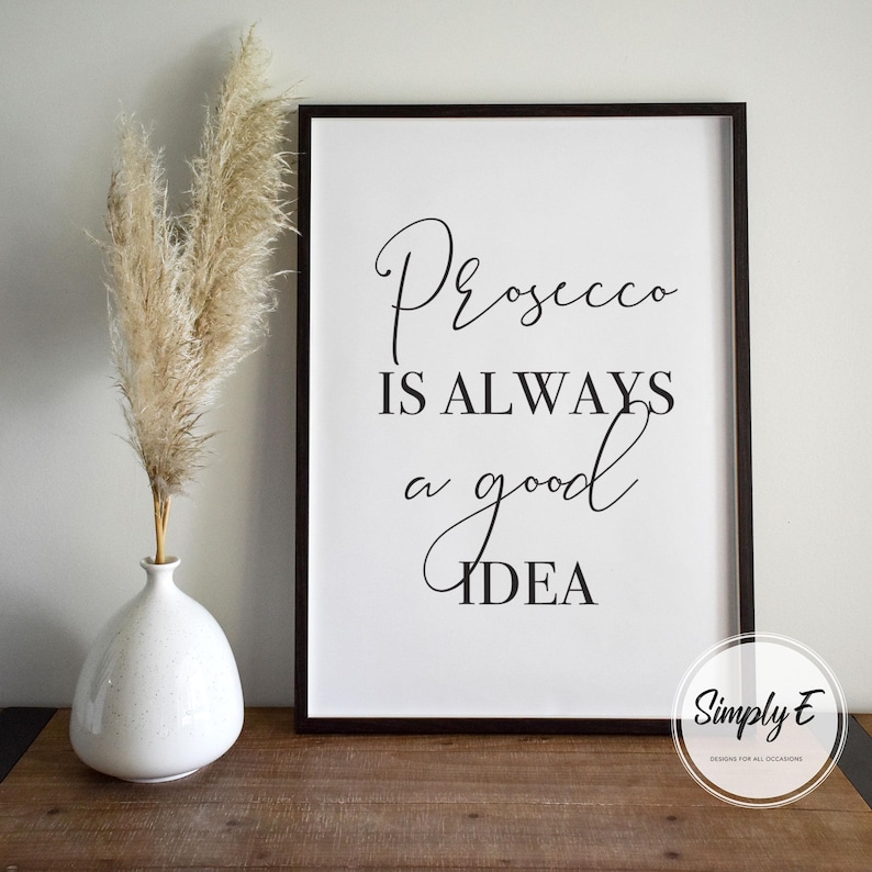 Op de afbeelding: Zwart-witte printbare wandkunst met de tekst "Prosecco is always a good idea". De print is ingelijst in een zwart frame en ligt op een houten oppervlak.