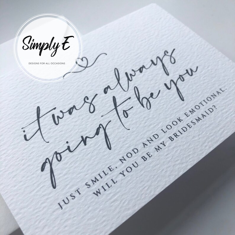 May include: A white card with grey text that reads "it was always going to be you" and "JUST SMILE, NOD AND LOOK EMOTIONAL. WILL YOU BE MY BRIDESMAID?"  The card has a small heart symbol above the text.