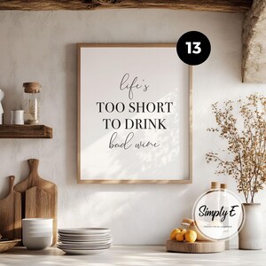 May include: A rectangular wall art print in a light wood frame featuring the black text Life's TOO SHORT TO DRINK bad wine. The print is displayed on a wall above a kitchen counter with wooden cutting boards, a stack of white plates, and a vase.