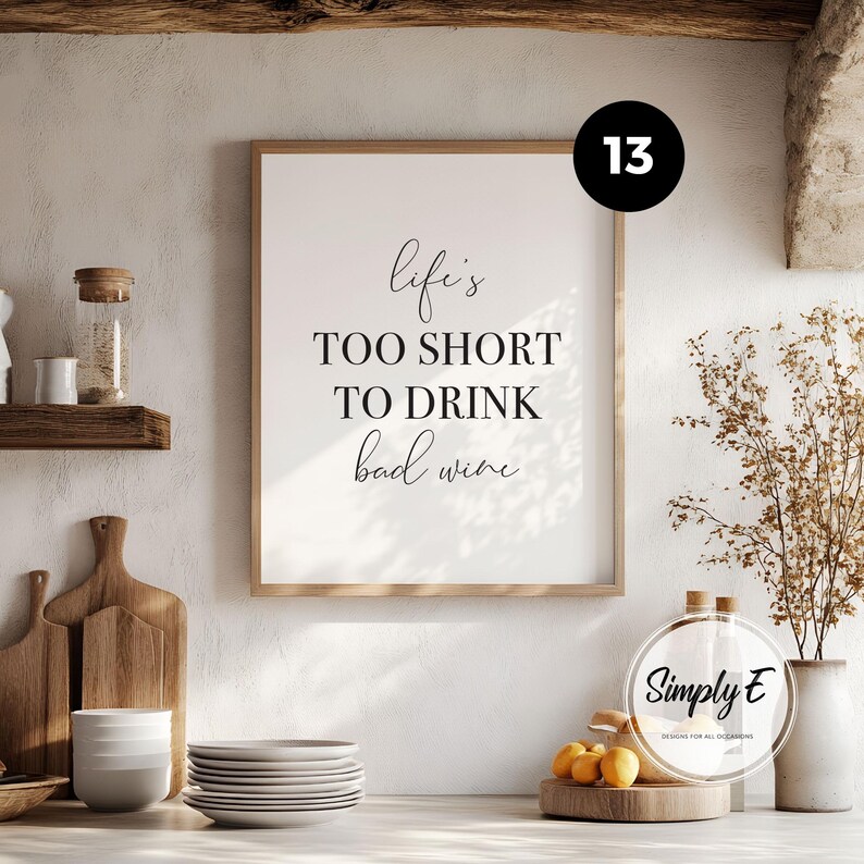 May include: A rectangular wall art print in a light wood frame featuring the black text Life's TOO SHORT TO DRINK bad wine. The print is displayed on a wall above a kitchen counter with wooden cutting boards, a stack of white plates, and a vase.