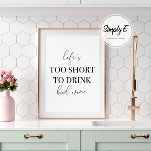 May include: A framed print with the text "Life's too short to drink bad wine" in black script. The print is in a white frame, set against a white and gray hexagonal tile backsplash. A pink vase with flowers and a gold faucet are also visible.