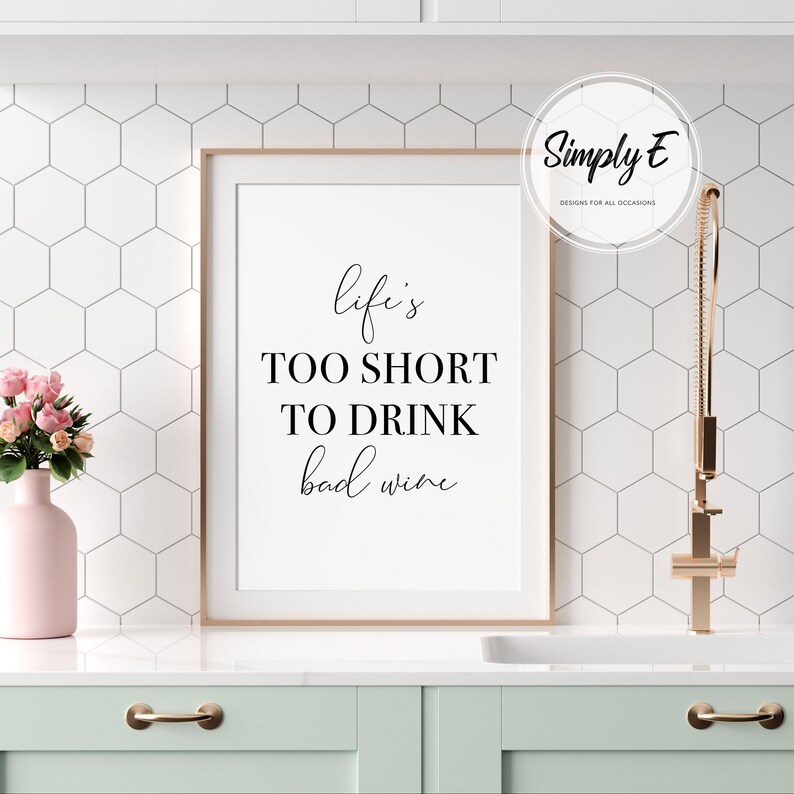May include: A framed print with the text "Life's too short to drink bad wine" in black script. The print is in a white frame, set against a white and gray hexagonal tile backsplash. A pink vase with flowers and a gold faucet are also visible.