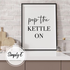 Peut inclure: Affiche murale imprimable en noir et blanc avec le texte "pop the kettle on" dans un style de calligraphie moderne. L'&oelig;uvre est encadr&eacute;e dans un cadre noir et est affich&eacute;e sur un mur en briques blanches.