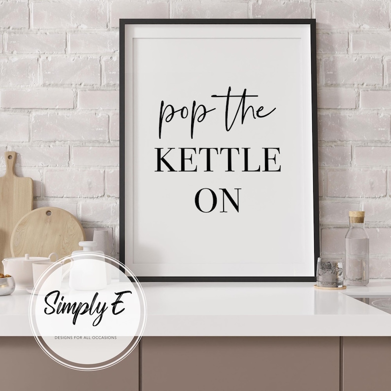 Peut inclure: Affiche murale imprimable en noir et blanc avec le texte "pop the kettle on" dans un style de calligraphie moderne. L'&oelig;uvre est encadr&eacute;e dans un cadre noir et est affich&eacute;e sur un mur en briques blanches.