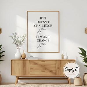 Może przedstawiać: Oprawiony wydruk z tekstem "IF IT DOESN'T CHALLENGE you IT WON'T CHANGE you" w czarnym skrypcie. Wydruk jest wyświetlany nad drewnianą szafką z trzema szufladami i dwiema drzwiami. Ozdobne wazony i rośliny znajdują się na szafce.