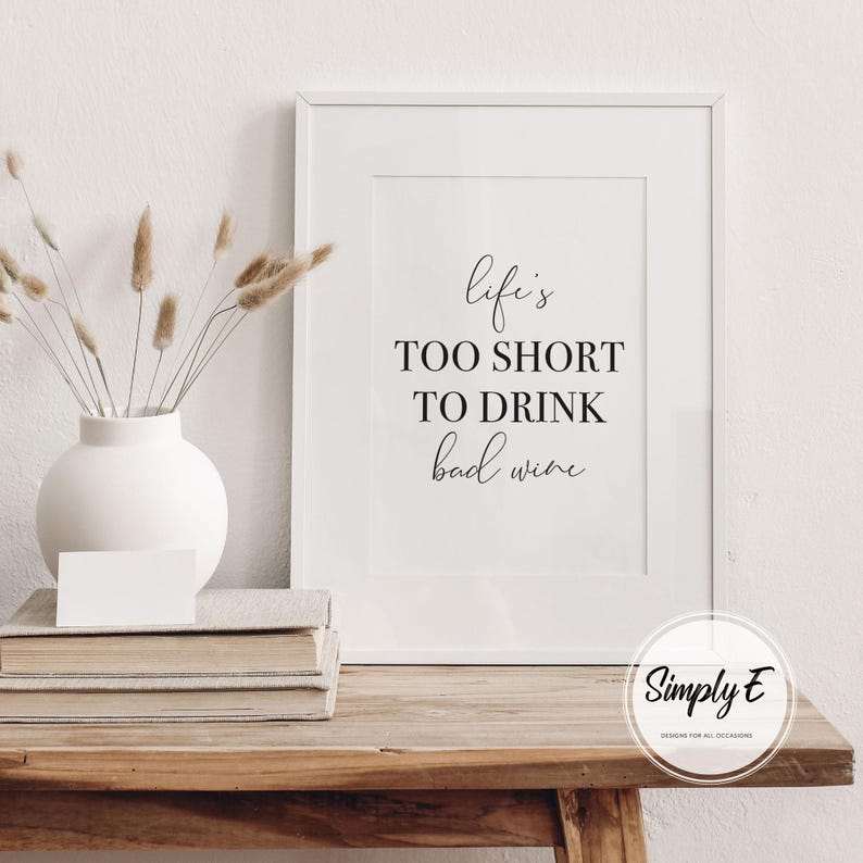 May include: A framed print with the text "Life's too short to drink bad wine" in black script. The print is displayed on a wooden table with a white vase holding dried flowers and a stack of books. A white card rests on the books.