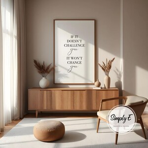 Może przedstawiać: Oprawiony wydruk z cytatem "If it doesn't challenge you, it won't change you" czarnym pismem. Wydruk wisi nad drewnianą szafką z wazonami i suszonymi roślinami. Okrągła pufa i nowoczesne krzesło znajdują się na pierwszym planie.