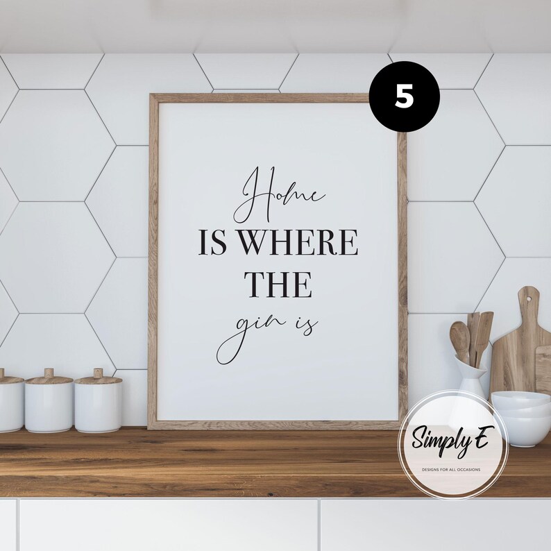 May include: A rectangular wall art print in a light wood frame featuring the black text Home IS WHERE THE gin is on a white background. The print is displayed on a wooden counter next to white kitchen canisters and wooden kitchen utensils.