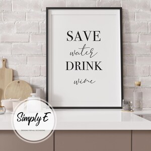 May include: A framed print with the text "SAVE water DRINK wine" in black lettering. The print is in a black frame and is displayed on a white countertop. The background is a white brick wall.