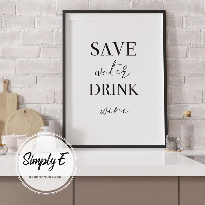 May include: A framed print with the text "SAVE water DRINK wine" in black lettering. The print is in a black frame and is displayed on a white countertop. The background is a white brick wall.
