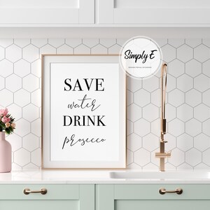 K&ouml;nnte beinhalten: Ein gerahmter Druck mit dem Text "SAVE water DRINK prosecco" in schwarzen Buchstaben auf wei&szlig;em Hintergrund. Der Rahmen ist in hellem Holz gehalten.
