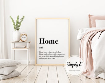 May include: A framed print with the definition of "Home" in black text. The print is in a gold frame and is propped against a wall. A small side table with a vase, clock, and books is to the left. A bed with white linens and pink pillows is to the right.