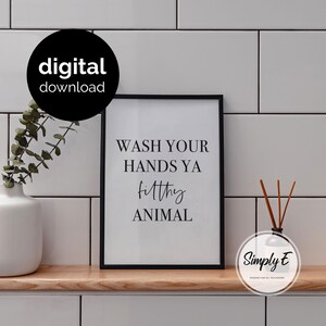 May include: A black framed wall art print featuring the white text WASH YOUR HANDS YA filthy ANIMAL in a mix of serif and script fonts. The print is displayed on a wooden shelf next to a white vase with greenery and a reed diffuser bottle.