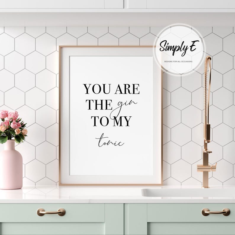 Puede incluir: Un estampado enmarcado con el texto "YOU ARE THE gin TO MY tonic" en escritura negra. El estampado est&aacute; en un marco blanco, sobre una pared de azulejos blancos y grises. Un jarr&oacute;n rosa con flores y un grifo dorado est&aacute;n en primer plano.