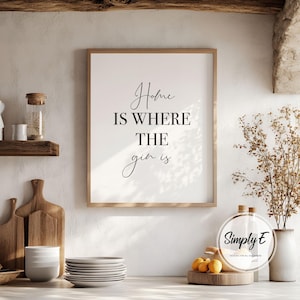 May include: A framed print with the text "Home is where the gin is" in a kitchen setting. The print has a wooden frame and is displayed above a countertop with plates, cutting boards, and decorative items. The overall aesthetic is rustic and inviting.
