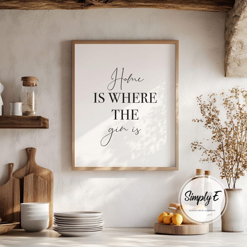 May include: A framed print with the text "Home is where the gin is" in a kitchen setting. The print has a wooden frame and is displayed above a countertop with plates, cutting boards, and decorative items. The overall aesthetic is rustic and inviting.