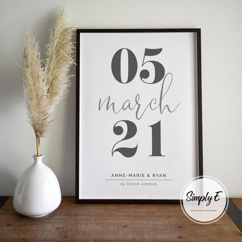 May include: A framed print with the date "05 march 21" in gray, with the names "Anne-Marie & Ryan" and address below. A white vase with dried pampas grass is to the left. The print is in a black frame.