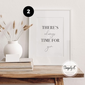 May include: A white framed wall art print featuring the black text THERE'S always TIME FOR gin. The print is displayed next to a white vase containing dried beige grass stems, resting on a stack of books. A black circle with the number 2 is visible.