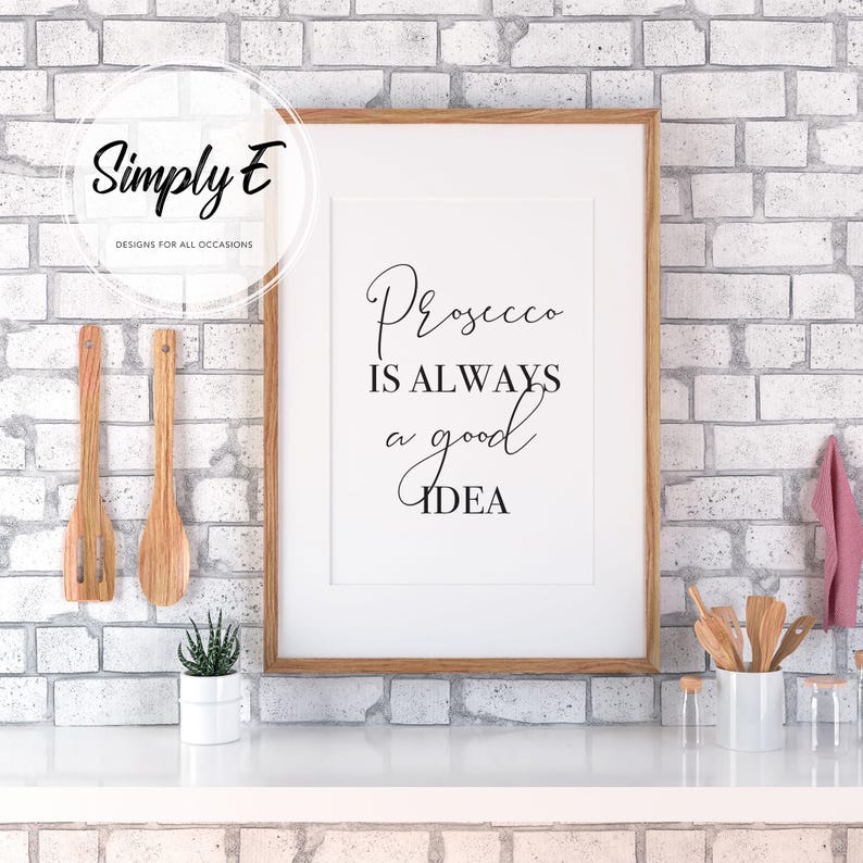 Op de afbeelding: Een ingelijst print met een witte achtergrond en zwarte tekst die "Prosecco is always a good idea" luidt. De print hangt aan een witte bakstenen muur in een keukenomgeving.