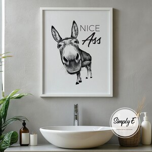 May include: Black and white framed art print featuring a donkey illustration with the text "NICE Ass". The artwork is displayed above a white sink in a bathroom setting. The print is in a white frame.