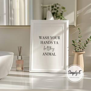 Puede incluir: Un marco blanco con el texto negro "WASH YOUR HANDS YA filthy ANIMAL". La impresi&oacute;n est&aacute; sobre una superficie blanca con un difusor, un jarr&oacute;n con vegetaci&oacute;n y un espejo en el fondo. La impresi&oacute;n es de Simply E Designs.