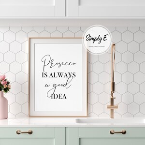 Op de afbeelding: Een witte ingelijste print met zwarte tekst die "Prosecco is always a good idea" luidt. De print hangt aan een witte muur met een wit zeshoekig tegelpatroon. Een ros&eacute;gouden kraan is zichtbaar op de achtergrond.