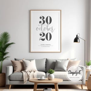 May include: A framed print with the date "30 October 20" in a stylish font. The print also includes the names "Jayne & Martin" and address "7 Ladybank Drive". The print is displayed above a gray sofa with decorative pillows.