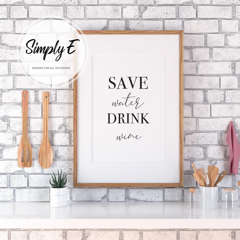 May include: A framed print with the text "SAVE water DRINK wine" in a modern font. The print is in a wooden frame and is displayed on a white surface against a white brick wall. Kitchen decor.