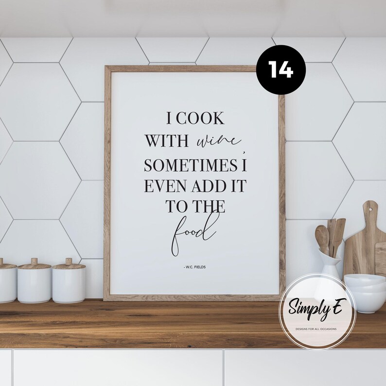 May include: A rectangular wall art print in a light wood frame featuring black text on a white background that reads: I COOK WITH WINE, SOMETIMES I EVEN ADD IT TO THE food, attributed to W.C. FIELDS. The print is displayed in a kitchen setting.