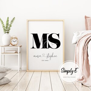 May include: A black and white printable wall art design with the letters "M&S" in a large font, with the names "marie & stephen" and the date "EST 2022" underneath. The design is framed in a gold frame and is hanging on a white wall.