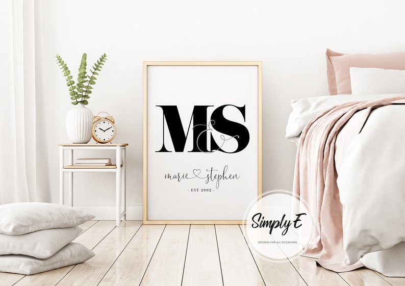 May include: A black and white printable wall art design with the letters "M&S" in a large font, with the names "marie & stephen" and the date "EST 2022" underneath. The design is framed in a gold frame and is hanging on a white wall.