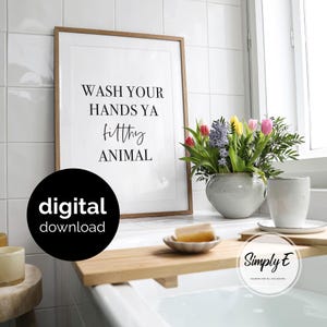 May include: A framed wall art print with a light wood frame featuring the black text WASH YOUR HANDS YA filthy ANIMAL on a white background. The print is displayed next to a ceramic vase with colourful tulips and a wooden bathtub tray.