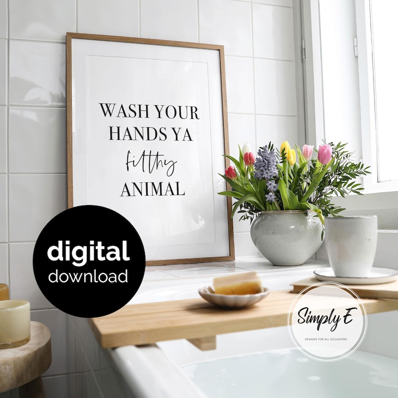 May include: A framed wall art print with a light wood frame featuring the black text WASH YOUR HANDS YA filthy ANIMAL on a white background. The print is displayed next to a ceramic vase with colourful tulips and a wooden bathtub tray.