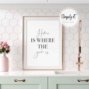 May include: A framed print with the text "Home is where the gin is" in a stylish script. The print is in a light wood frame and is displayed in a kitchen setting. A pink vase with flowers is on the counter.