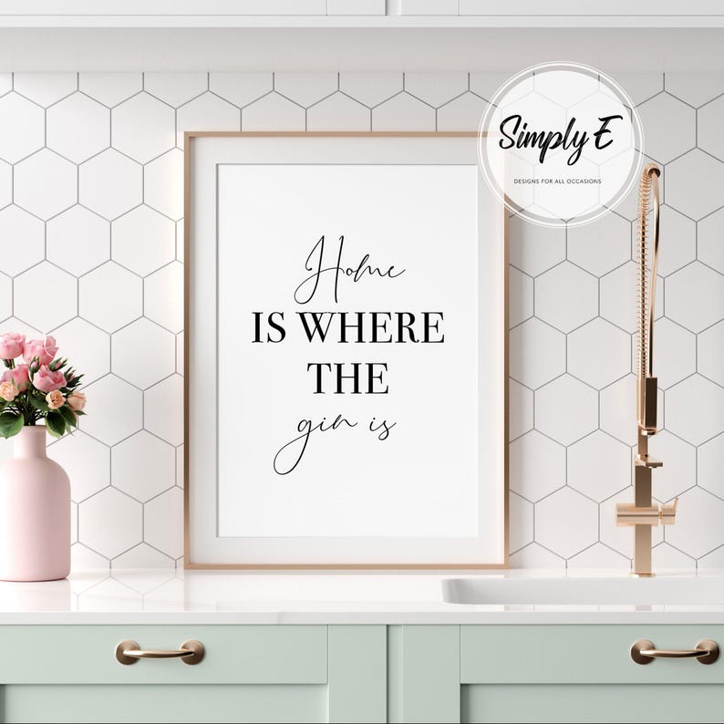 May include: A framed print with the text "Home is where the gin is" in a stylish script. The print is in a light wood frame and is displayed in a kitchen setting. A pink vase with flowers is on the counter.